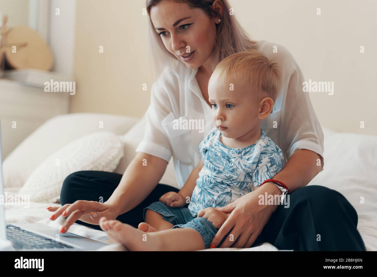 Young mother with her child working on laptop in bedroom at home. Multi-tasking, freelance and motherhood concept Stock Photo