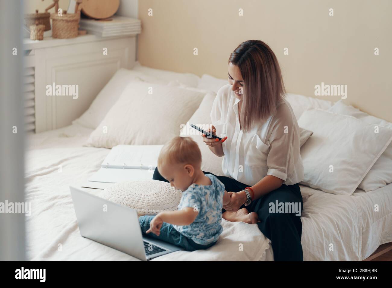 Young mother with her child working on laptop in bedroom at home. Multi-tasking, freelance and motherhood concept Stock Photo