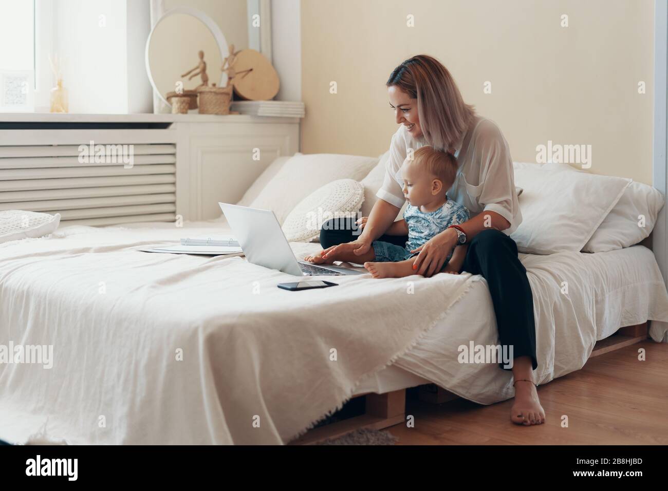 Young mother with her child working on laptop in bedroom at home. Multi-tasking, freelance and motherhood concept Stock Photo