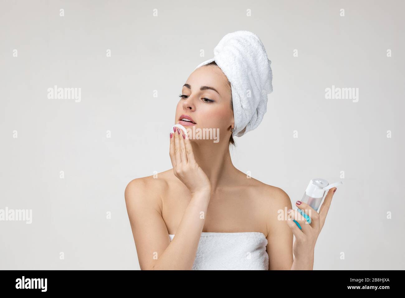 Young woman in towel removing makeup from her face with cotton pad