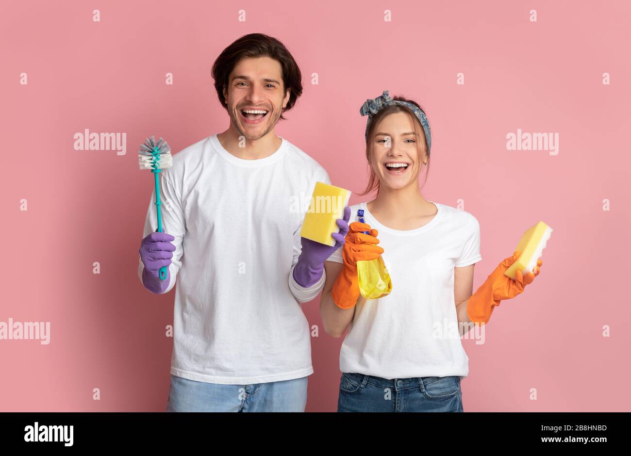 Excited couple wearing gloves, holding cleaning supplies in hands, pink background Stock Photo