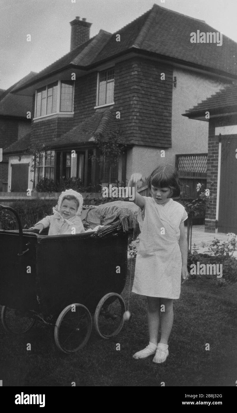 1940s, a young girl with her infant sister, who is sitting in a leather lined pram outside in a front garden on a suburban street, England, UK. Stock Photo