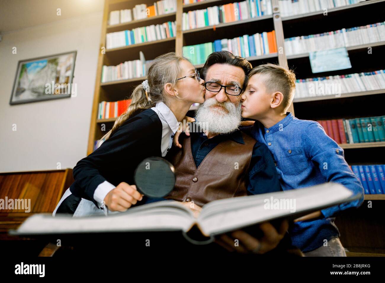 Lovely portrait of happy children, boy and girl, kissing their old bearded granddad in cheeks while spending time, reading amazing book together in Stock Photo