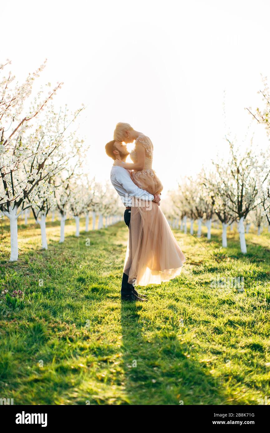 Charming tender loving couple walking in a spring blossoming garden at sunrise. Man whirls woman in her arms. Love story, wedding concept, romantic Stock Photo