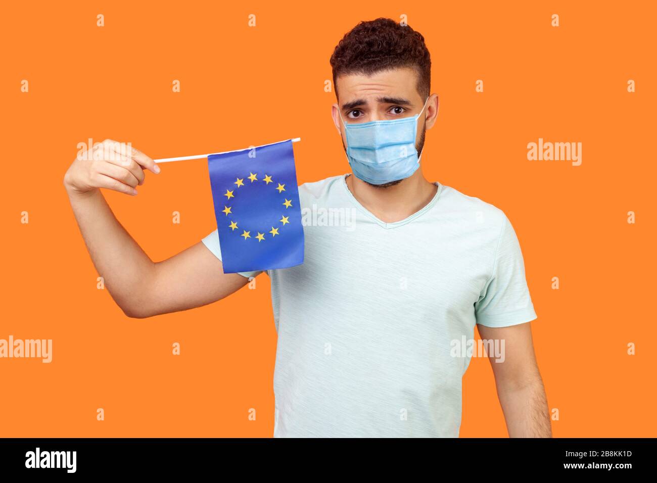 European Union flag. Portrait of upset man with medical mask in white t-shirt holding EU flag and looking at camera with disappointed sad expression. Stock Photo