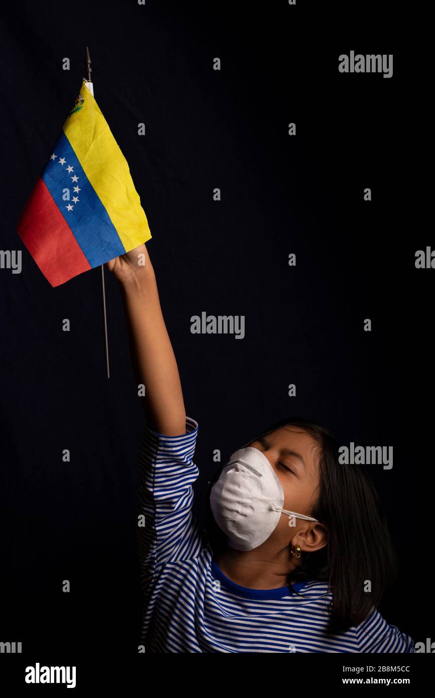 Portrait of little Venezuelan girl wearing medical mask and holding hopefully the flag of Venezuela Stock Photo