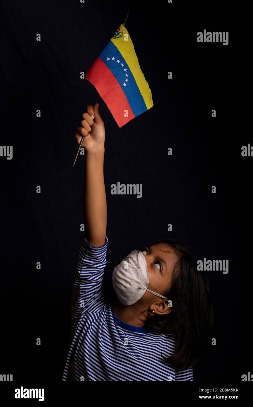 Portrait of little Venezuelan girl wearing medical mask and holding hopefully the flag of Venezuela Stock Photo