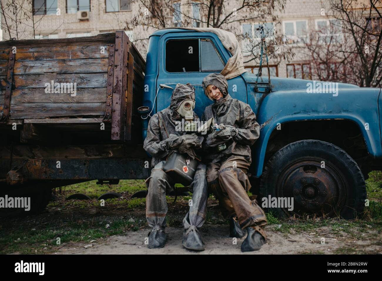 Couple in love sits in NBC protective suits and gas mask near old truck. Concept of a preventive measures and protection for coronavirus COVID 19 pand Stock Photo