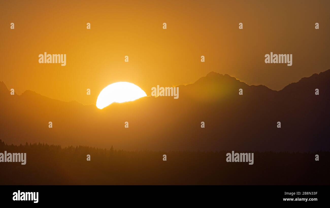 The sun peeks behind the Olympic Mountains as it sets for the evening casting a golden glow on the sky and mountain range Stock Photo