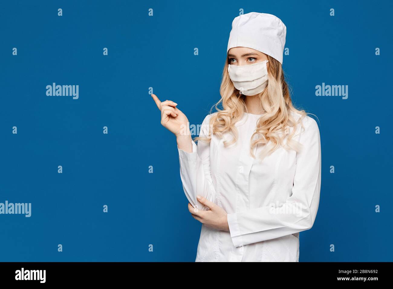 Young woman in medical uniform and face mask posing at blue background ...