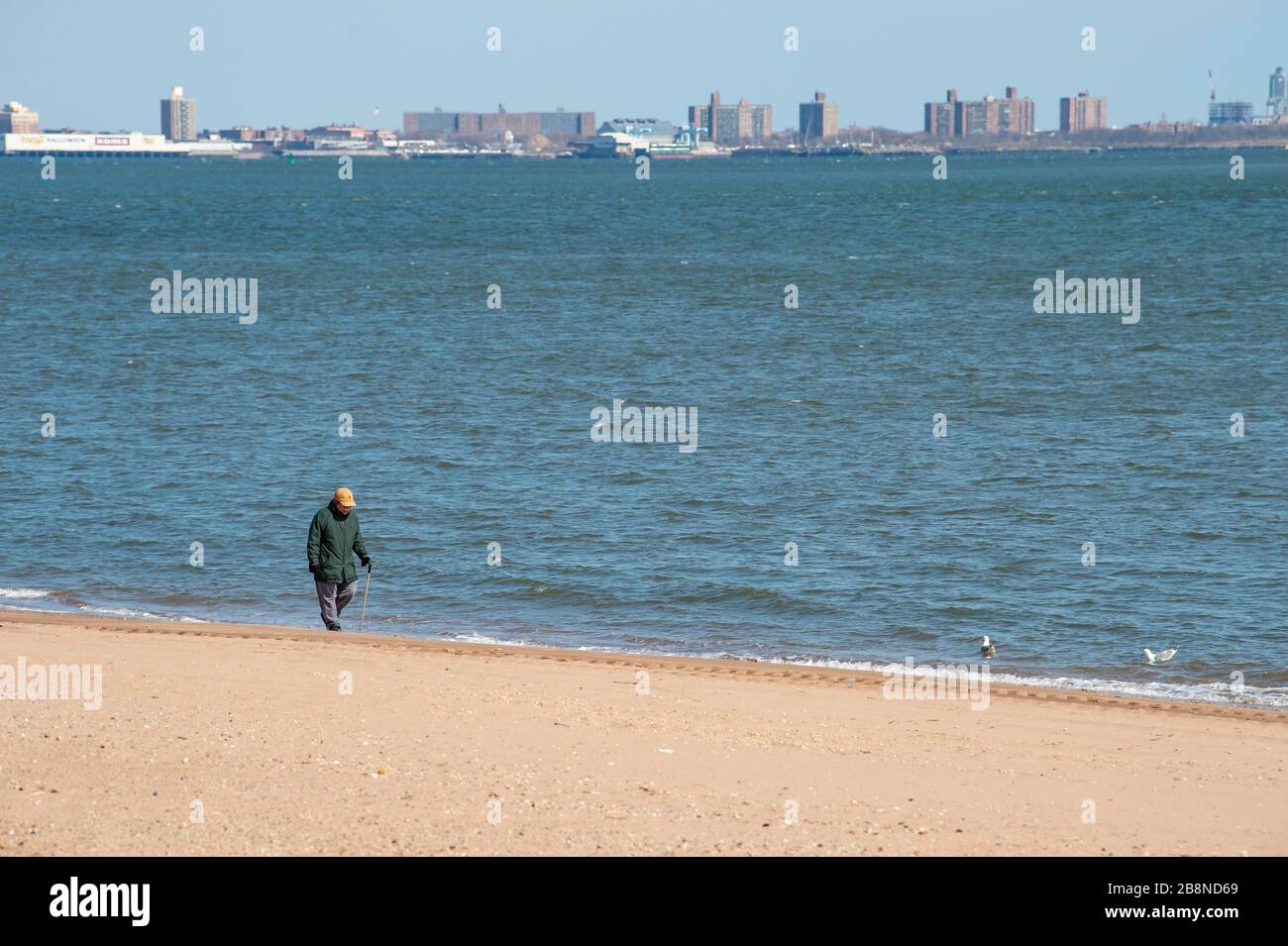 Staten Island, New York, USA. 22nd Mar, 2020. A man walks alone on the beach with Brooklyn in the background in Staten Island, New York. Mandatory credit: Kostas Lymperopoulos/CSM/Alamy Live News Stock Photo