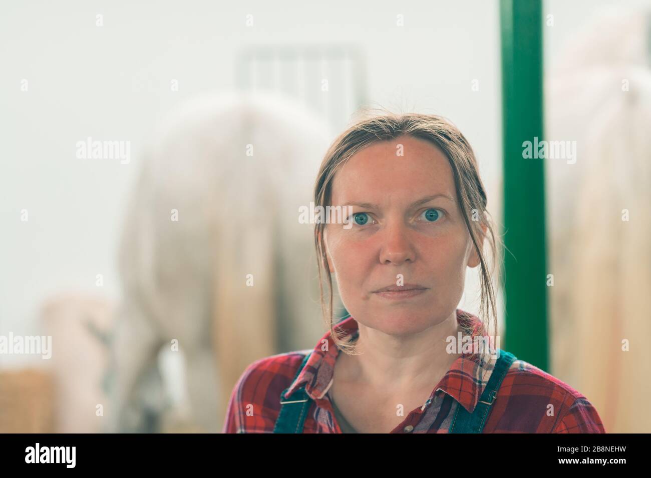 Portrait of female rancher at horse stable looking at camera. Adult woman wearing plaid shirt and jeans bib overalls as farm worker. Stock Photo