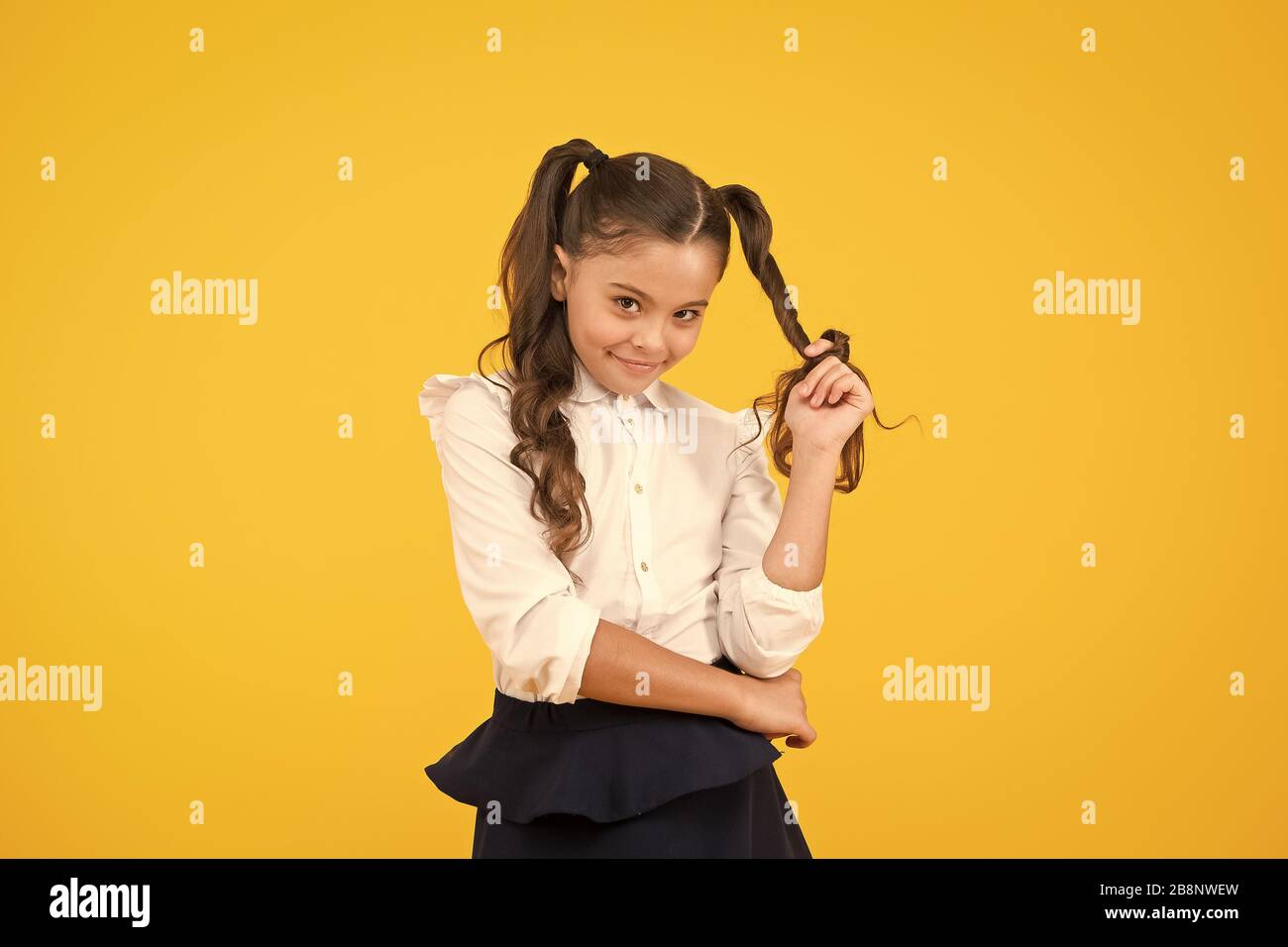 Modest hairstyle. Cute little schoolchild winding hairstyle around her finger on yellow background. Fashion girl with long ponytail hairstyle in formal style. Small kid styling hairstyle for school. Stock Photo