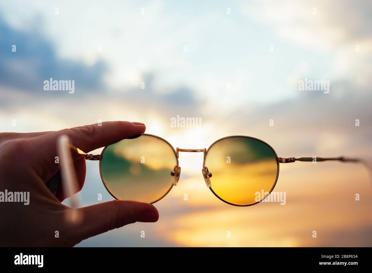 light skinned hand holds metal round sunglasses through which you can see a beautiful sea sunset in the mountains of blue and orange colors Stock Photo