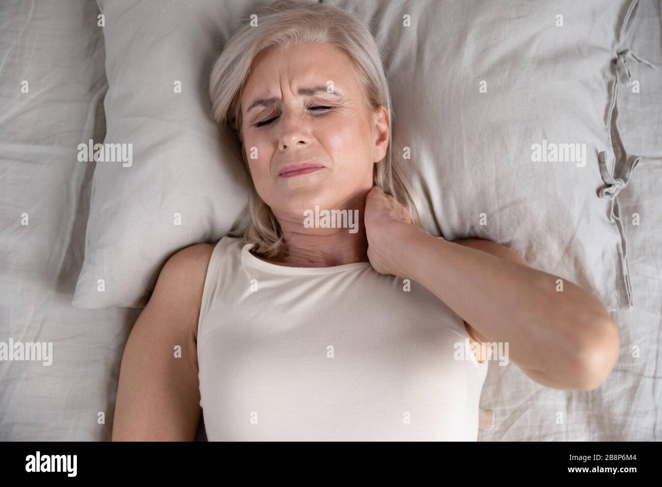 Older unhappy woman lying in bed, feeling strong neck ache. Stock Photo