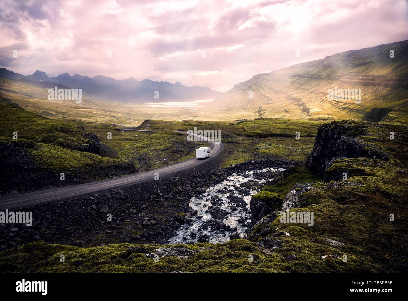 white camper van driving along winding mountain road through beautiful sunlit valley surrounded by mountains, Iceland Stock Photo