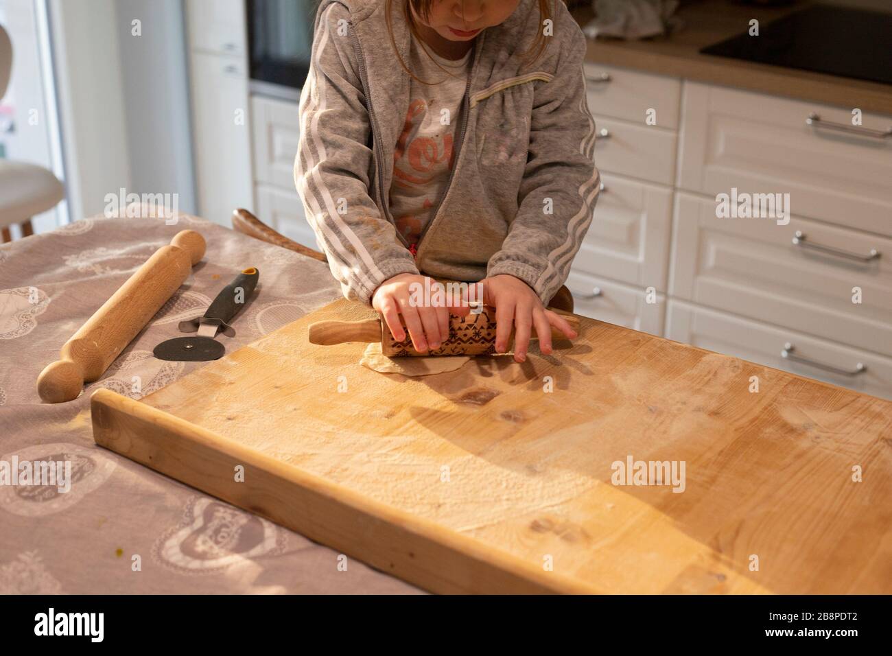 Child girl in white kitchen flattening pizza dough with a small rolling pin on a wooden board. Lockdown activity idea. Stock Photo