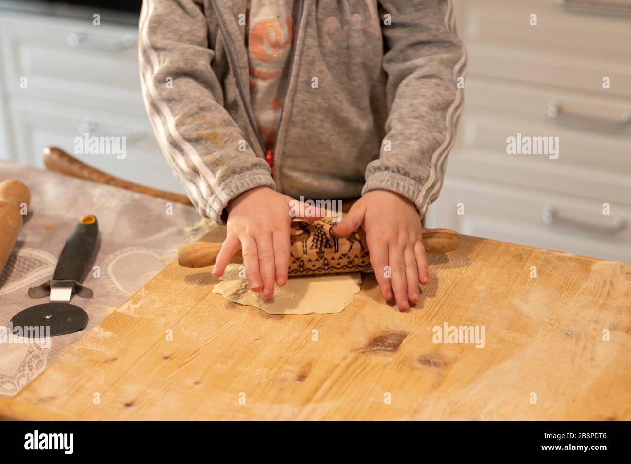 Child girl hands, in white kitchen, flattening pizza dough with a small rolling pin on a wooden board. Lockdown activity idea. Stock Photo