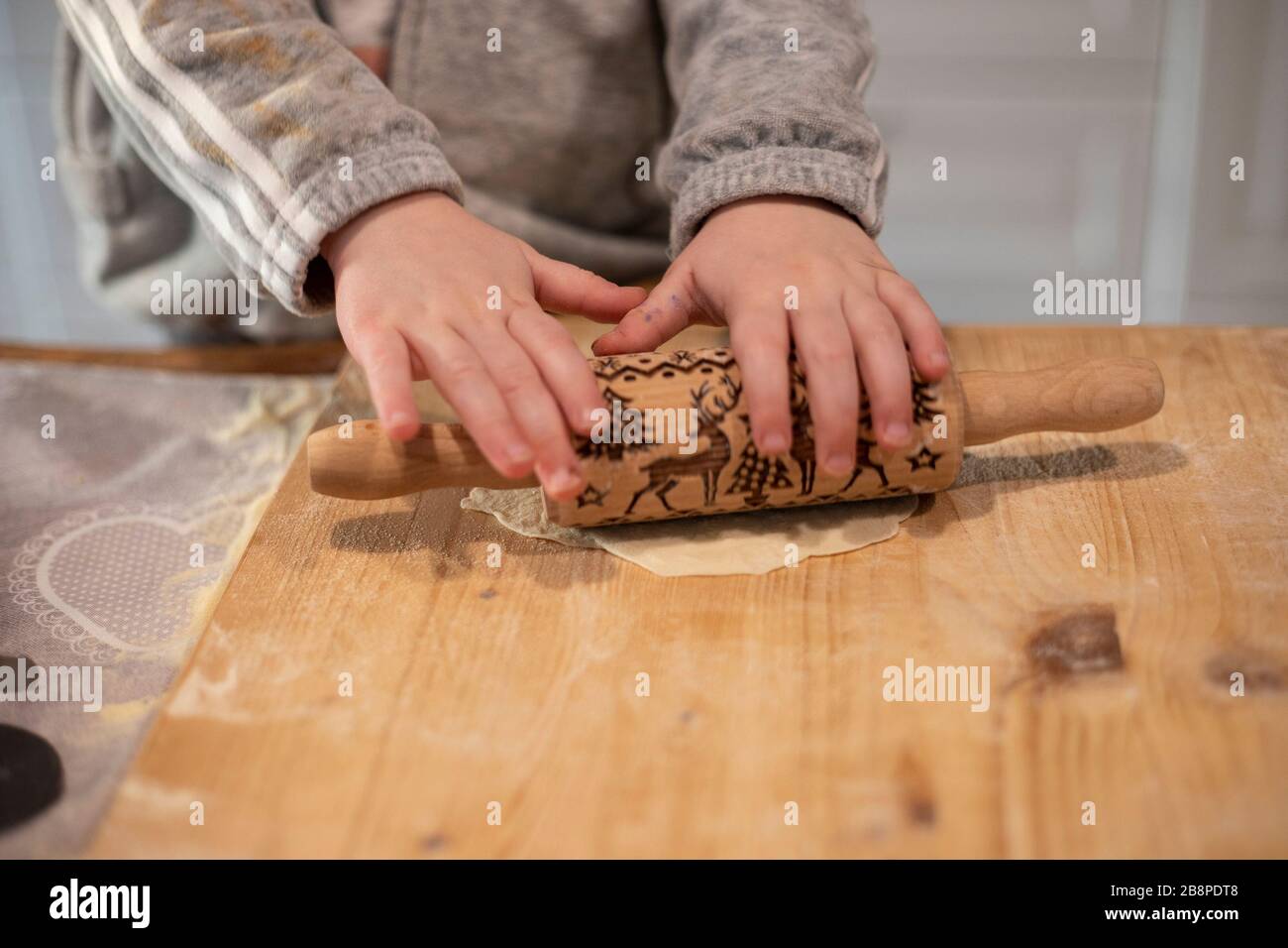 Child girl hands, in white kitchen, flattening pizza dough with a small rolling pin on a wooden board. Lockdown activity idea. Stock Photo