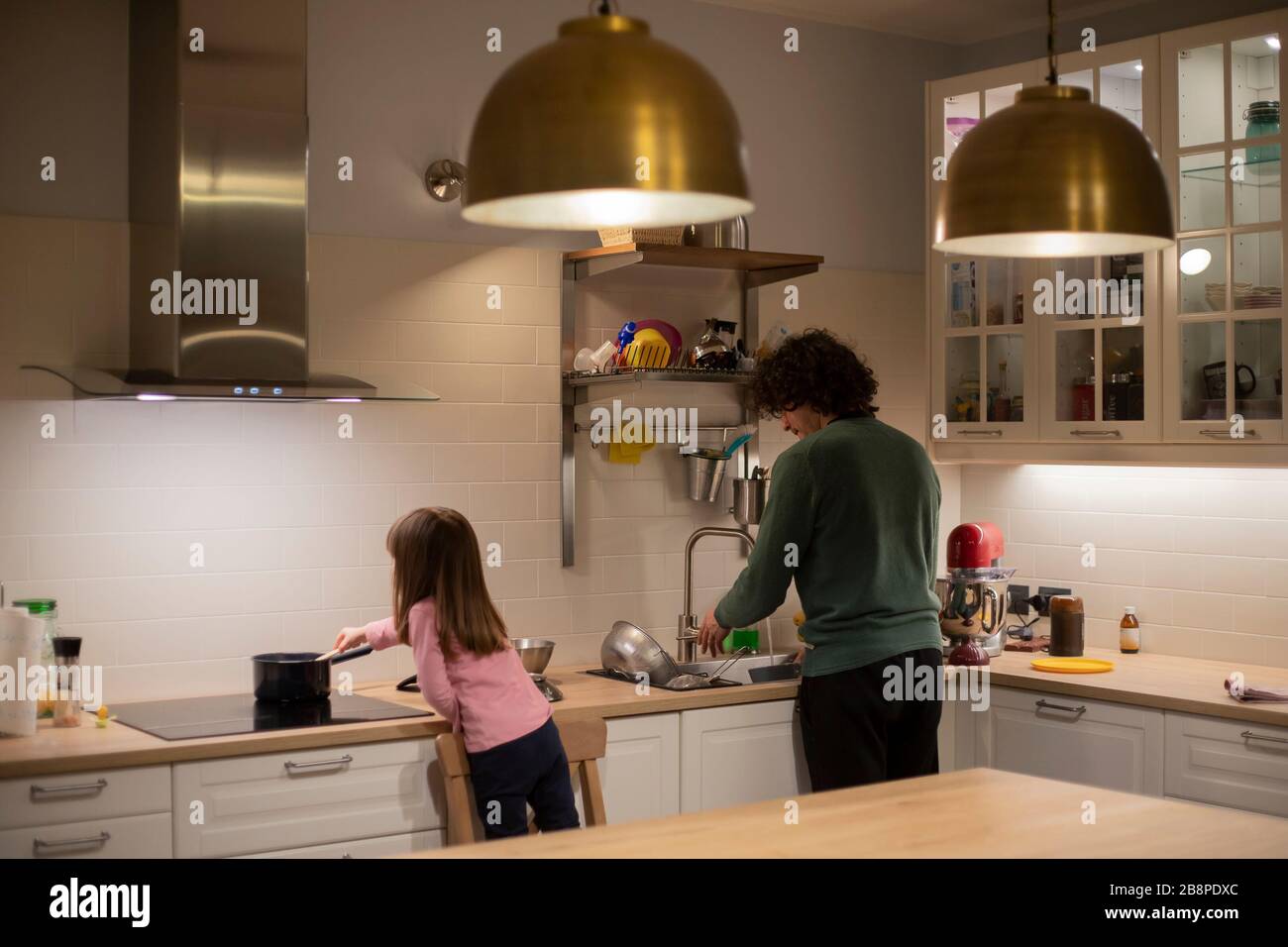 Young father and little child girl, back view, cooking together in modern, well lit white kitchen with big brass lamps. Lockdown activity idea. Stock Photo