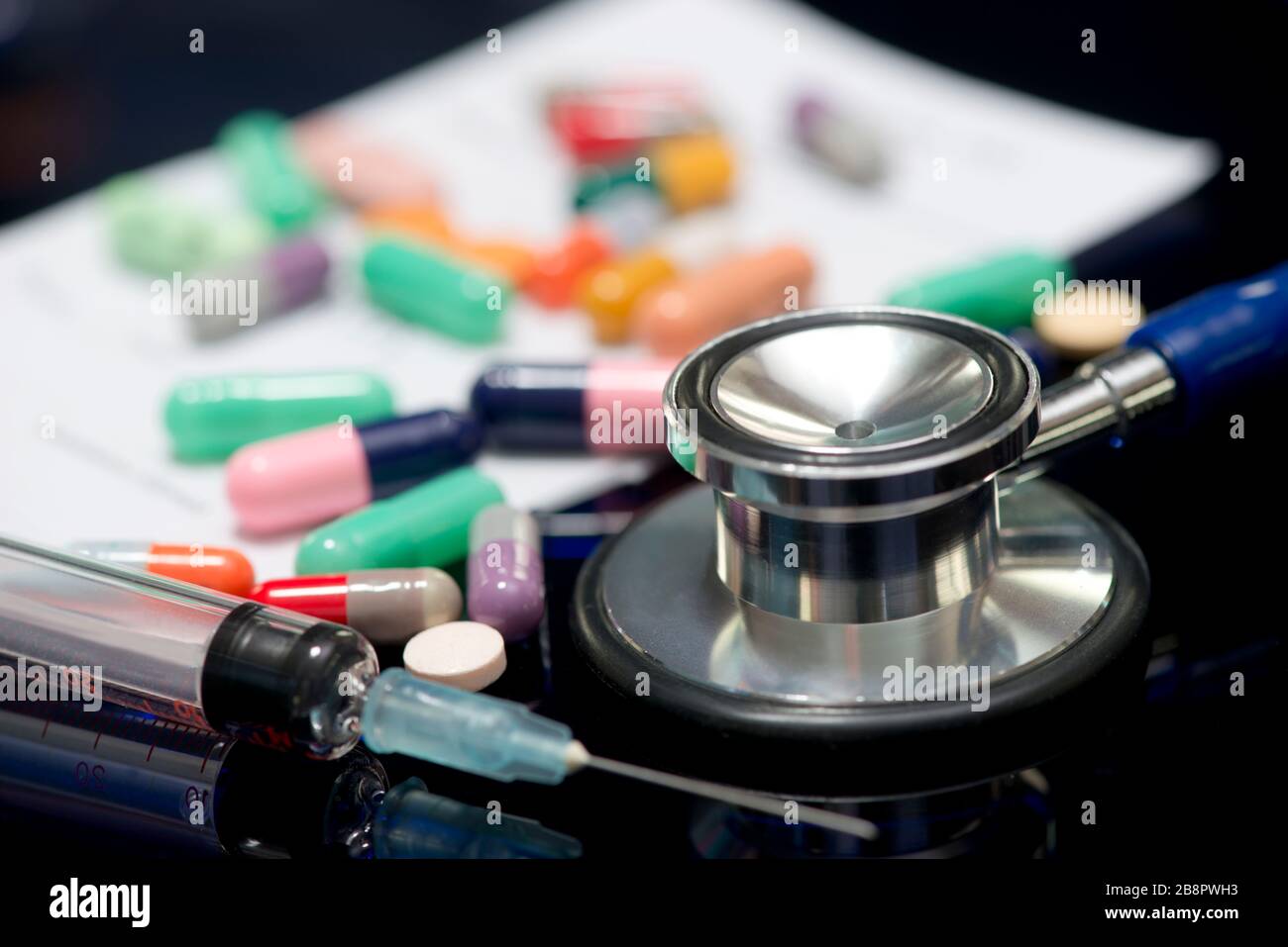 Stethoscope, syringe, different colored medications, and written doctor's prescription on dark reflective surface. Stock Photo