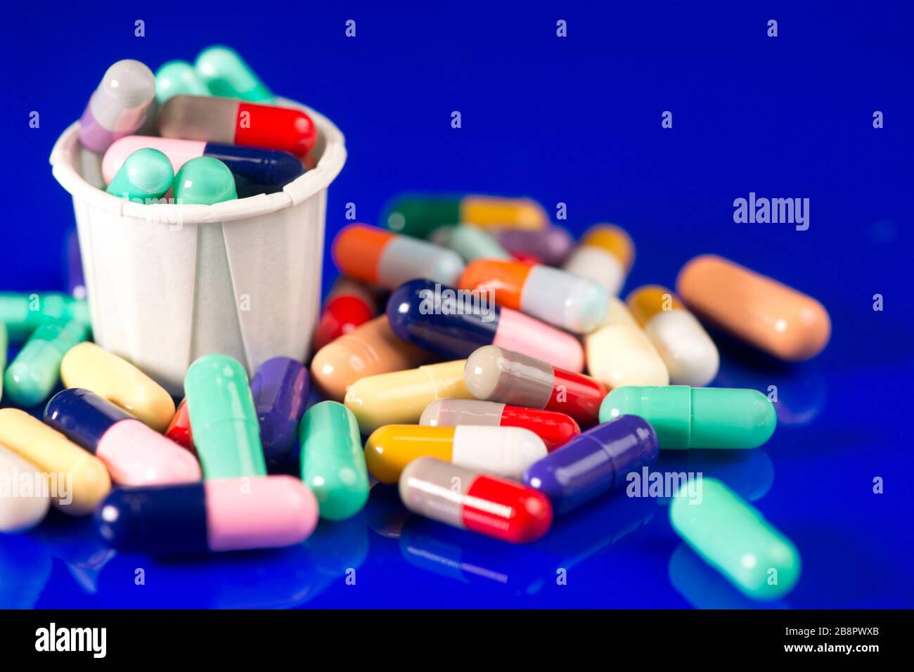 Assorted many colored capsules with paper medication cup on blue reflective surface. Stock Photo