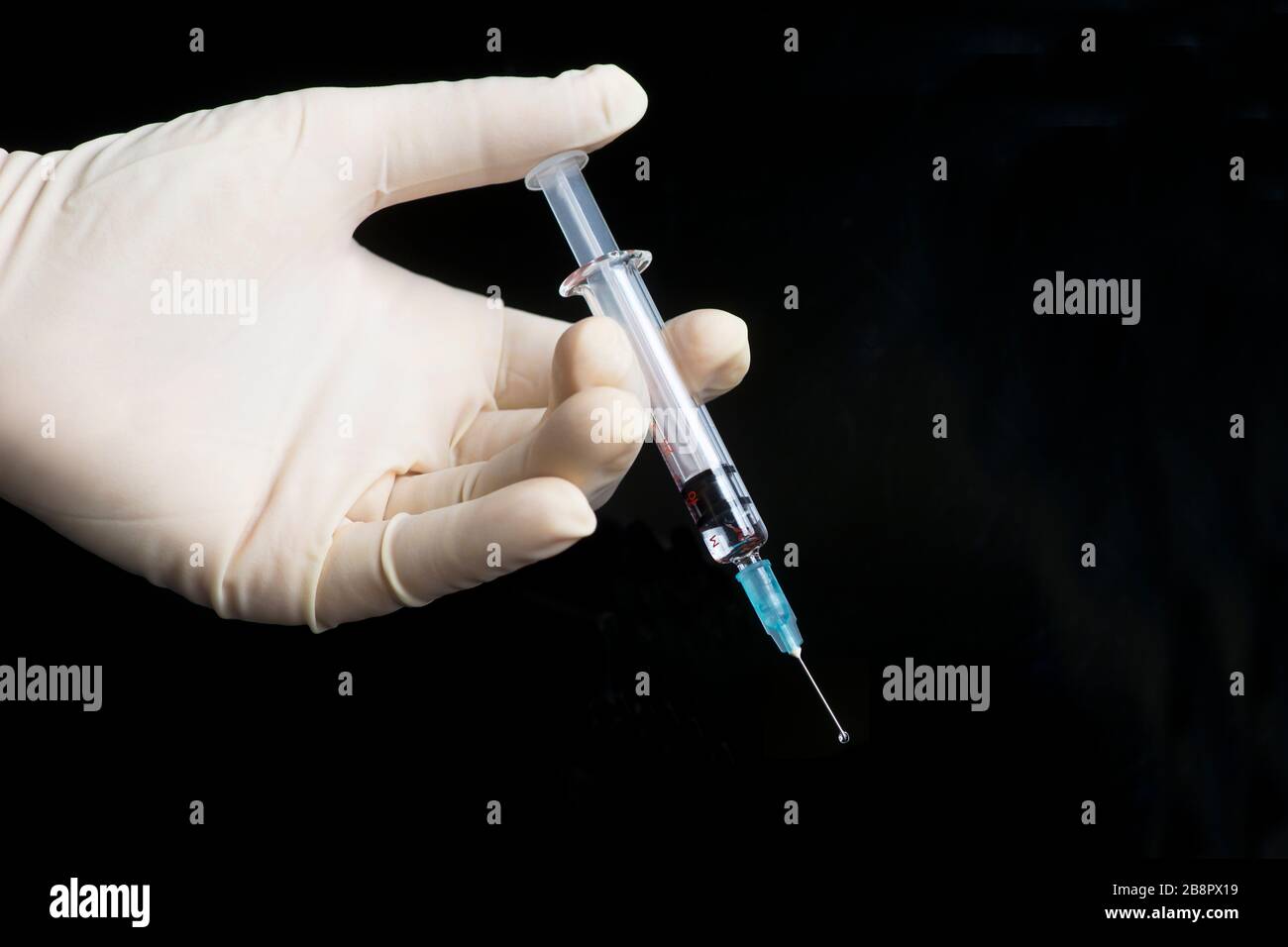 Gloved hand holds syringe with droplet on black background. Stock Photo
