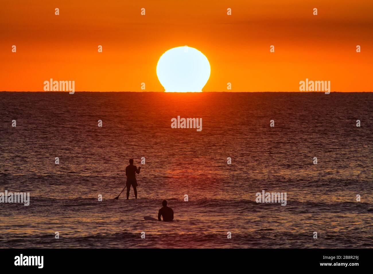 Two stand up paddleboarders facing an over sized sun at sunset on the ocean. Stock Photo