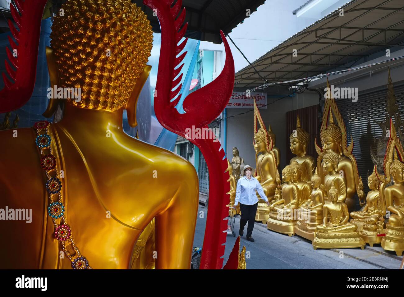 A woman passes through an alley off Bamrung Muang Rd., Bangkok, Thailand, past a row of Buddha statues manufactured in adjacent small factories Stock Photo