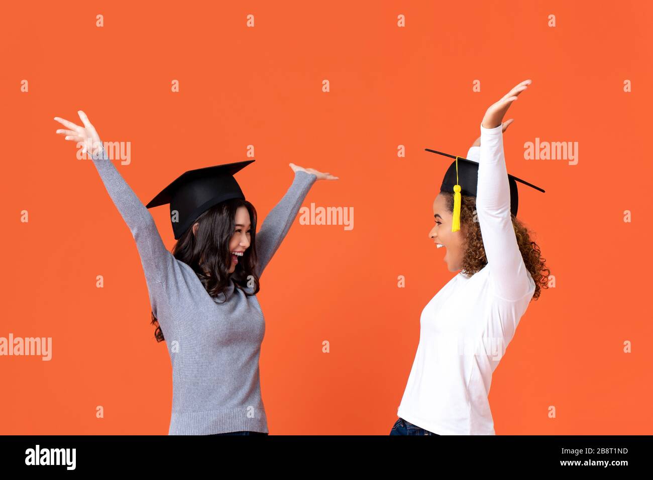 Happy excited young female students wearing graduate caps smiling with hands raising celebrating graduation day isolated on orange background Stock Photo