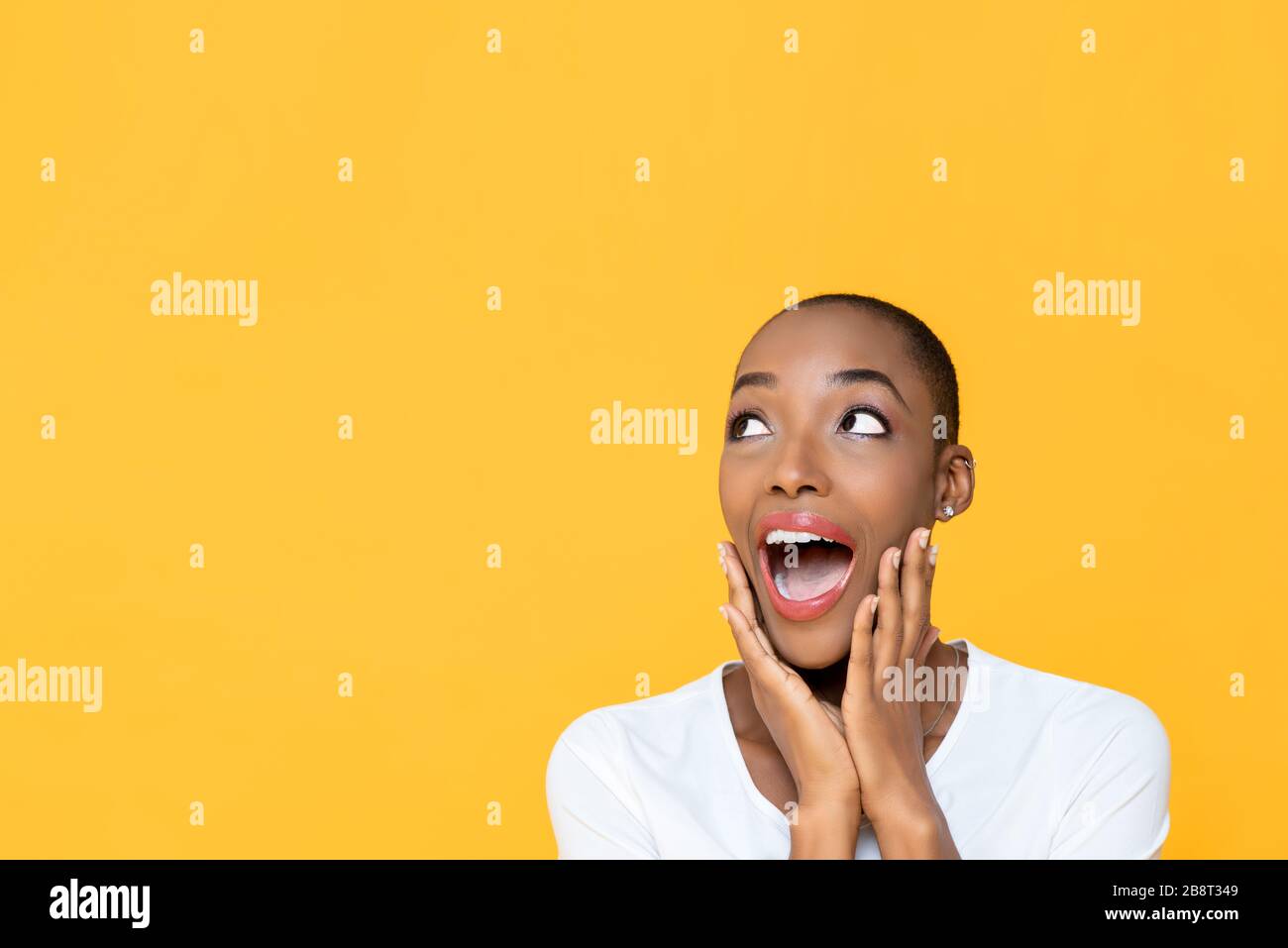 Shocked African American woman with hands on cheeks gasping and looking up to copy space aside isolated on yellow background Stock Photo