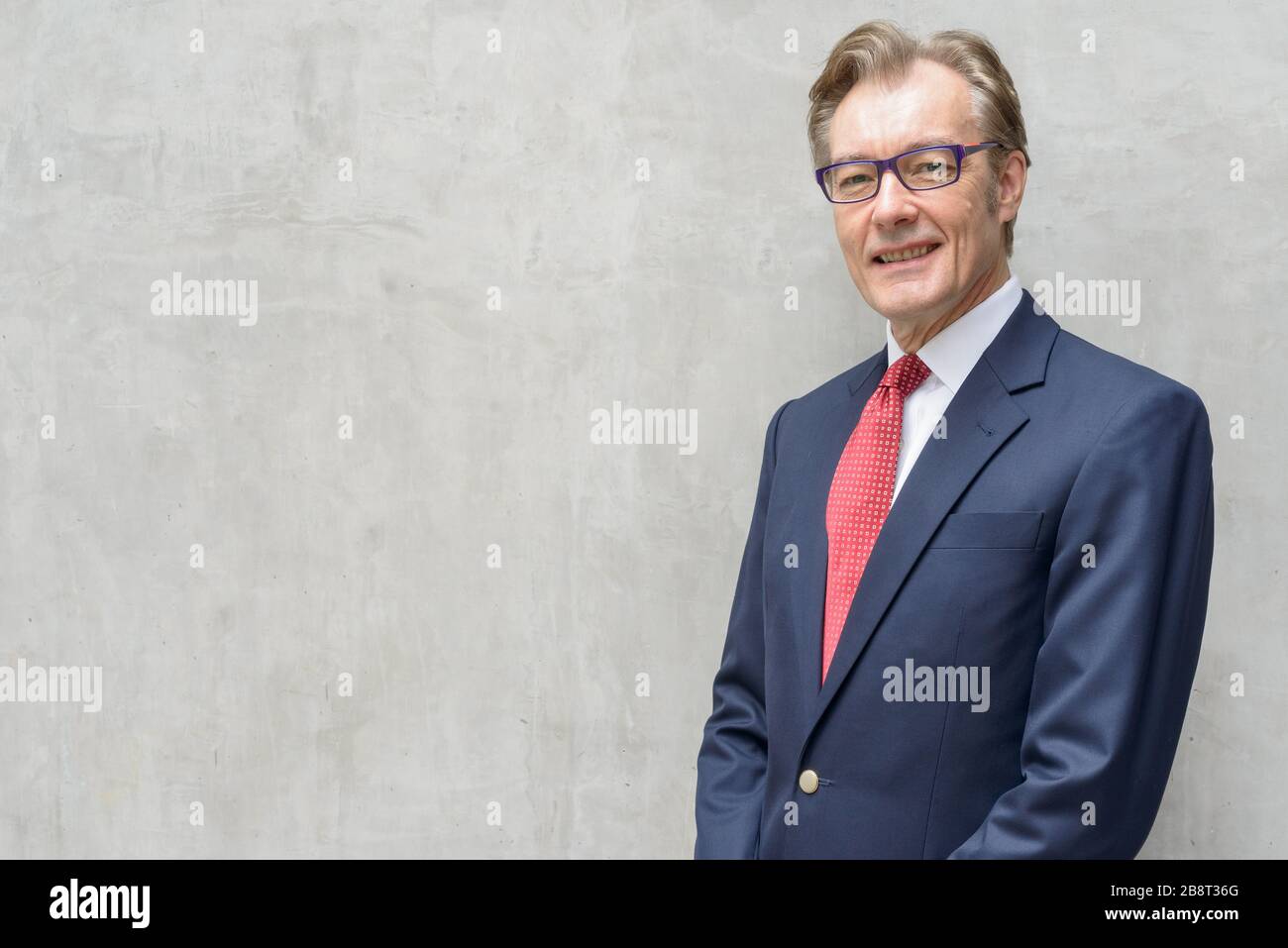 Happy handsome mature businessman in suit smiling against concrete wall ...