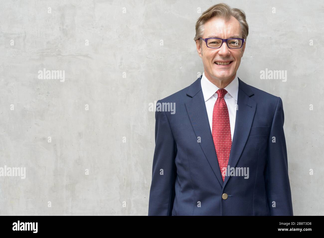 Happy handsome mature businessman in suit smiling against concrete wall ...
