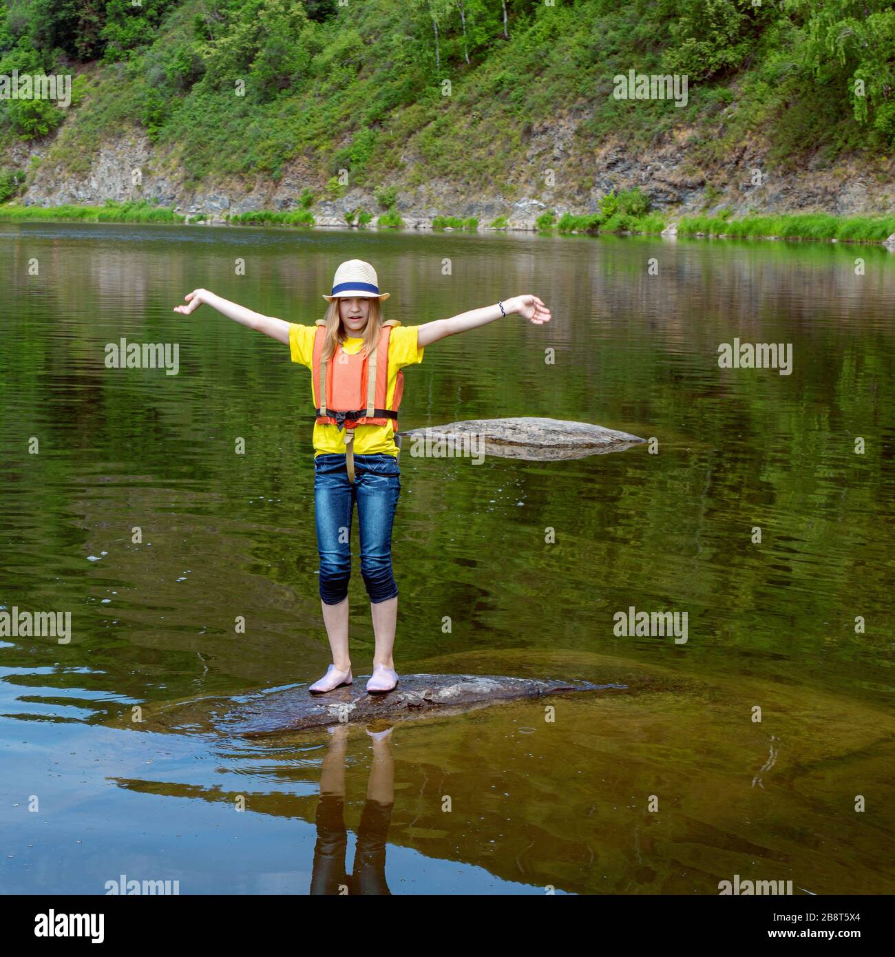 Young girl in life jacket standing on stone in middle of water. Waving hands, calling for help. Victim of shipwreck, accident. Stock Photo