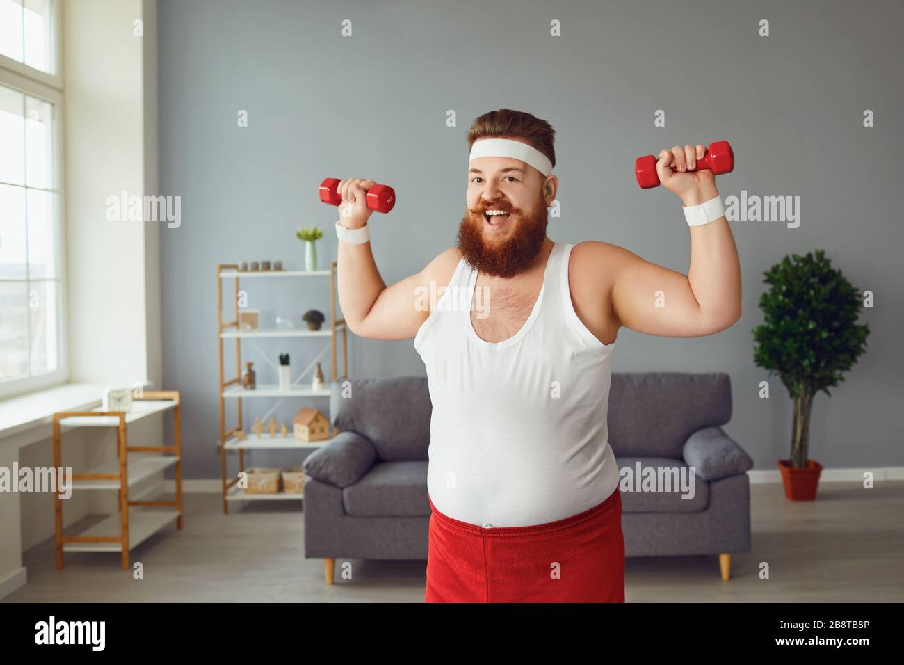 Funny fat man with dumbbells doing exercises in the room. Stock Photo