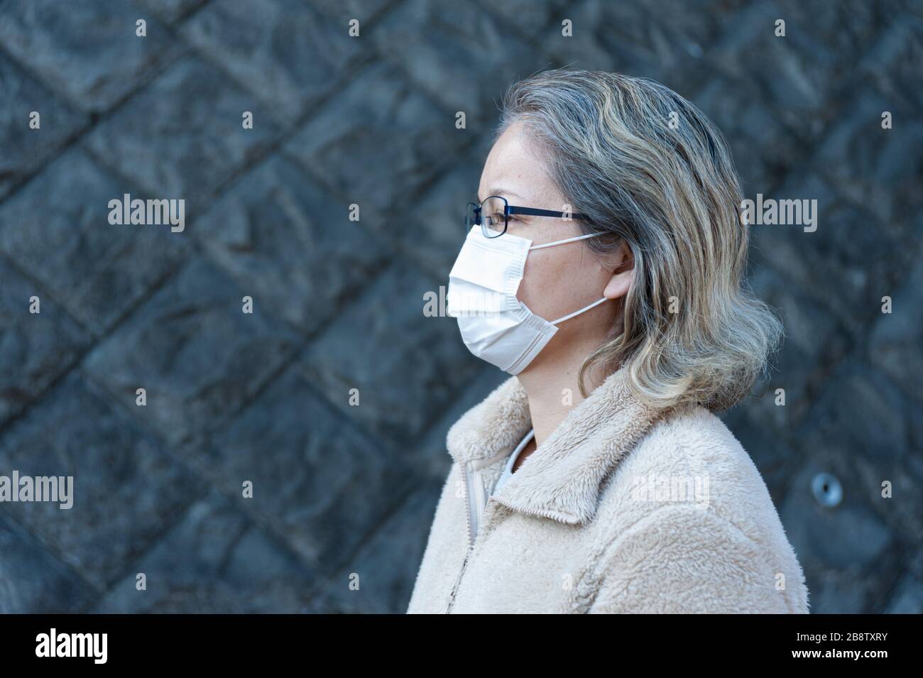 Woman 40-49 year old outdoors wearing glasses and white mask to protect against viruses, flu, hay fever and other diseases. Looking to the side. Stock Photo