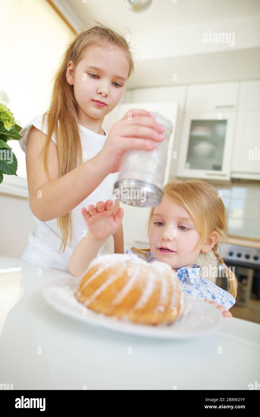Two children bake a cake in the kitchen and decorate it with icing sugar Stock Photo