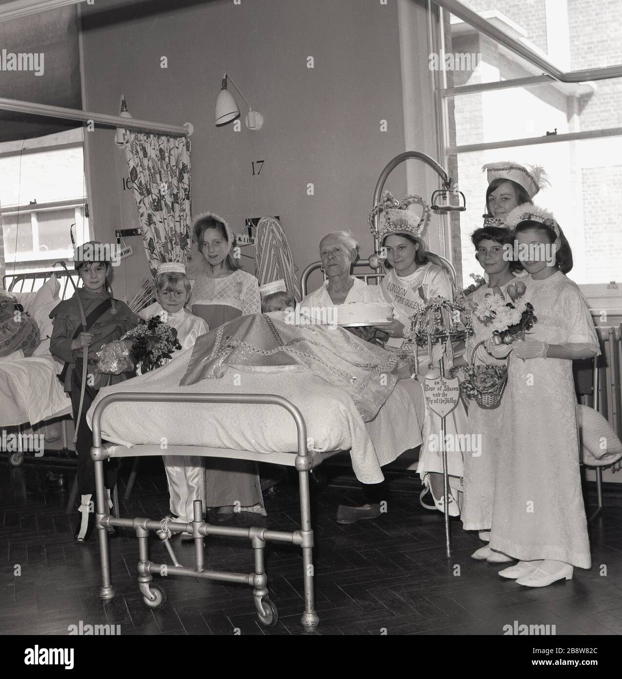 1960s, historical, a visit to a hospital ward by the newly crowned London May Queen,a young teenage girl and entourage, a group of young children dressed in costumes, Lewisham, South East London, England, UK. The children sit beside an elderly male patient sitting in his bed having presented him with a birthday cake. Stock Photo