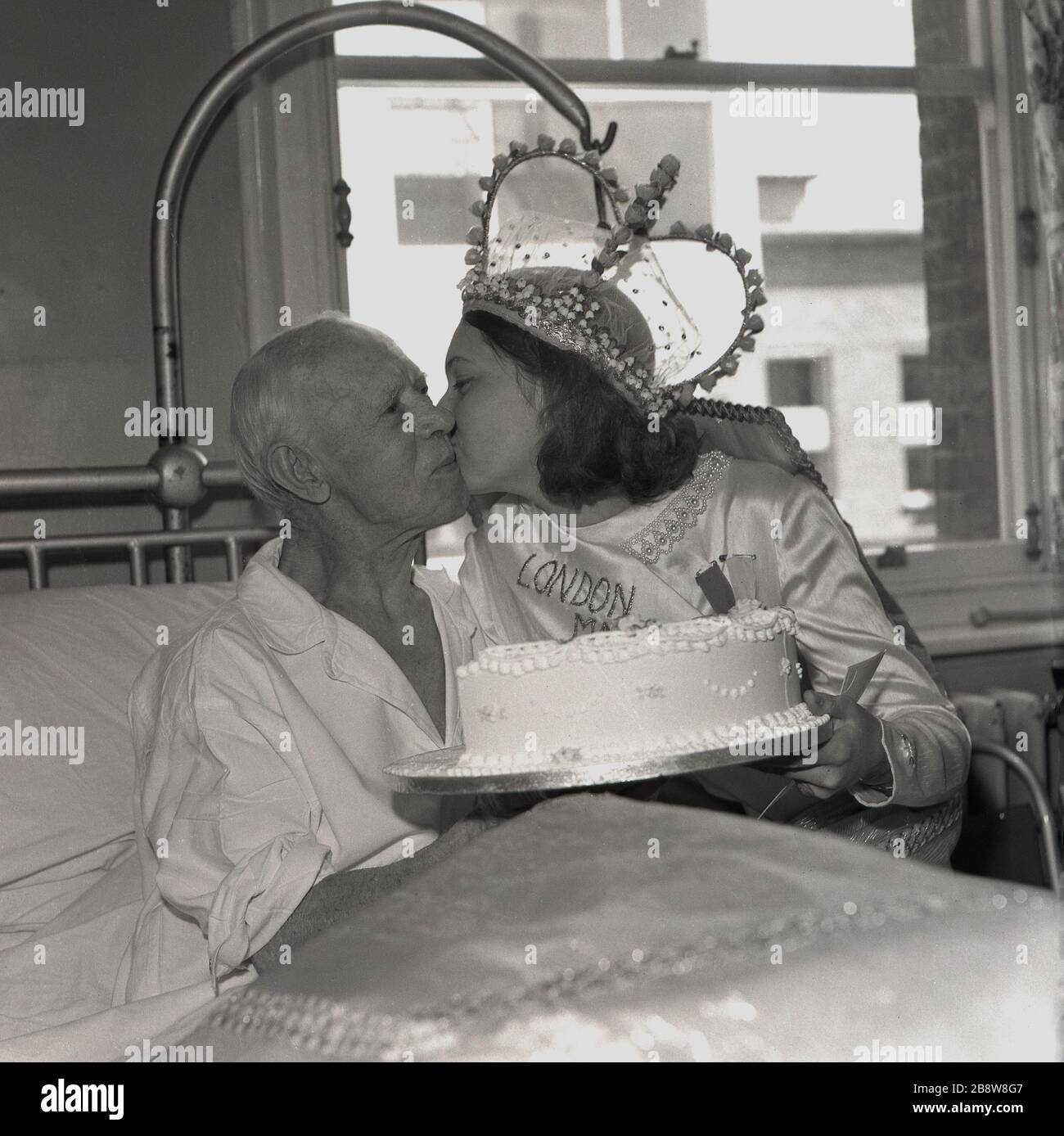 1960s, historical, a visit to a hospital by the London May Queen. The young teenage girl presents a cake and gives a kiss on the cheek to an elderly male patient sitting in his bed, Lewisham, South East London, England, UK. Stock Photo
