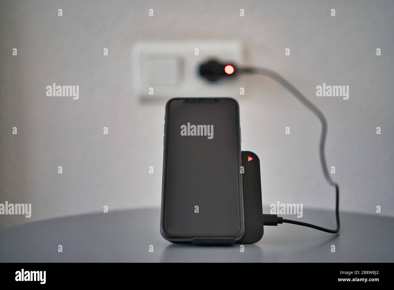 Black desktop wireless charger with charging led charging a smartphone. Display in black. Silver gray table. Plug at the wall. Stock Photo