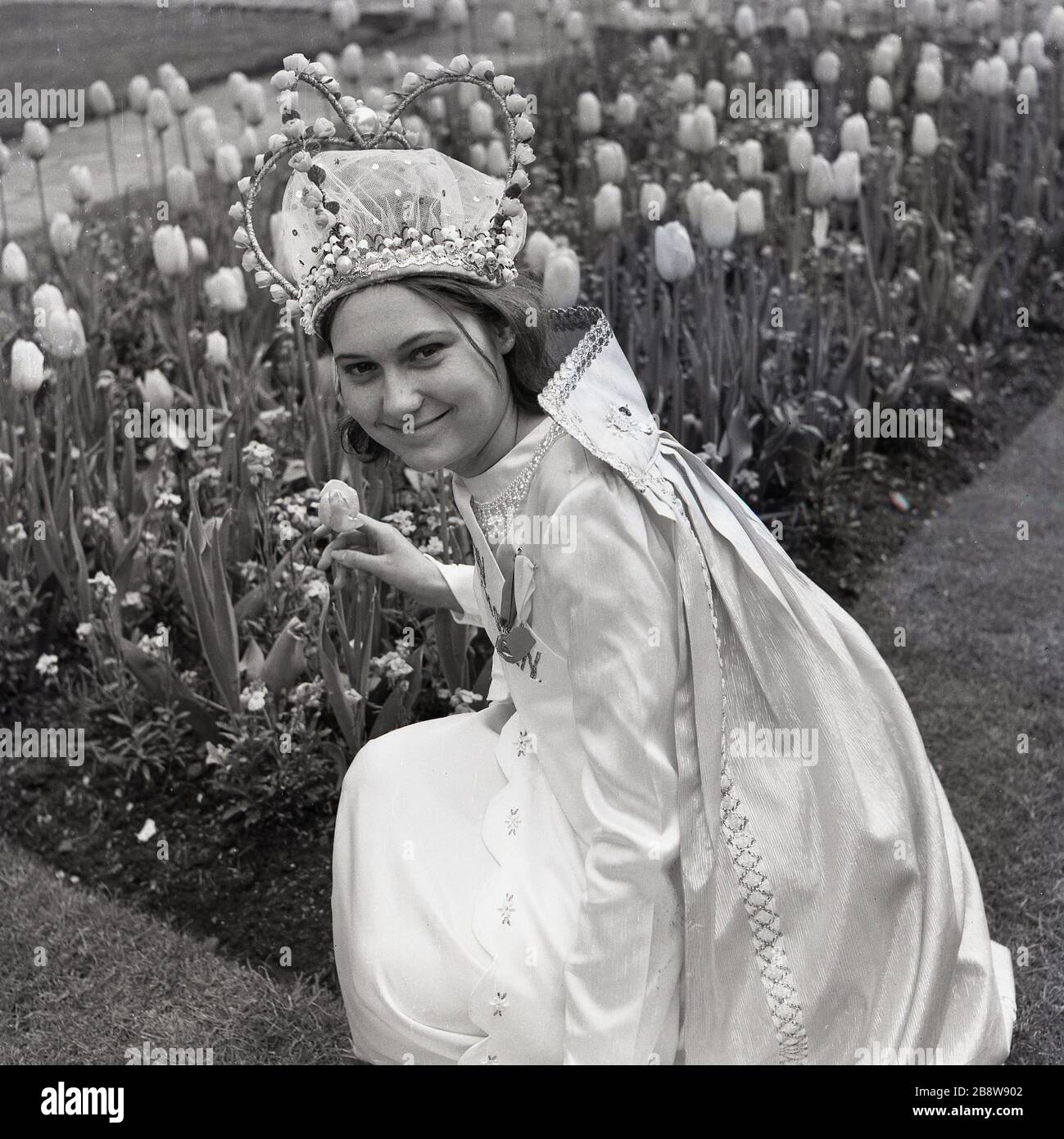 1960s, historical, the newly crowned London May Queen, a young teenage girl wearing her gown and tiara, posing for a picture outside by some flowers, Lewisham, South East London, England, UK. Stock Photo