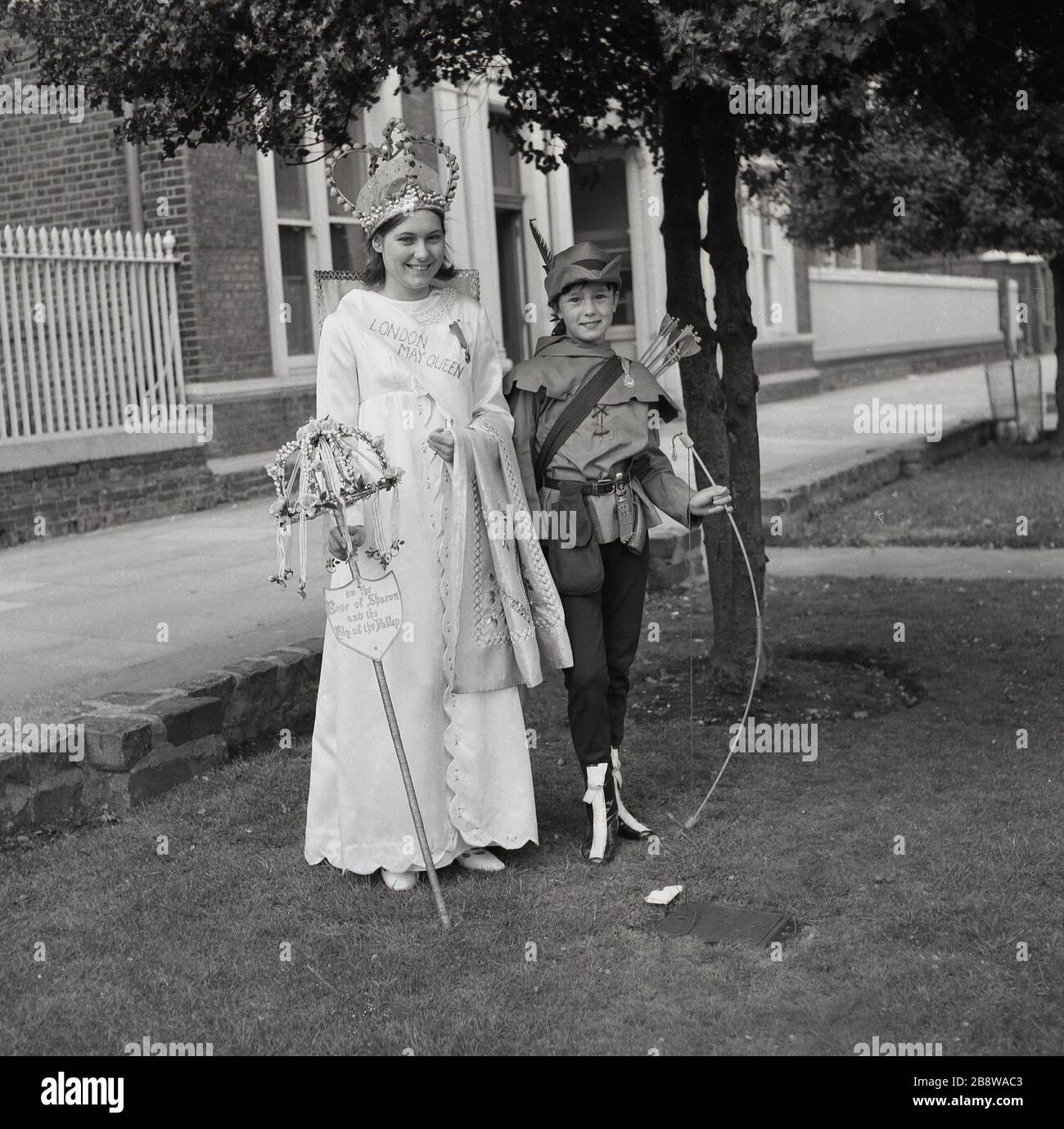 1960s, historical,a young teenage girl, newly crowned as the London May Queen in gown and tiara, standing beside a young girl in a costume holding a bow and arrow, Lewisham, South East London, England, UK. Stock Photo