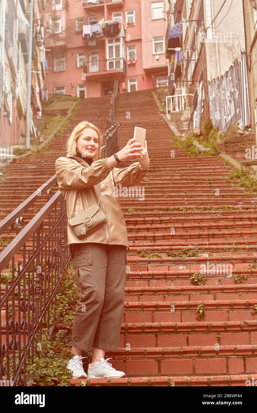 Young caucasian female traveler taking a selfie on the red stairs in Istanbul, using a smartphone, or mobile phone. Woman tourist travels to Turkey. Stock Photo