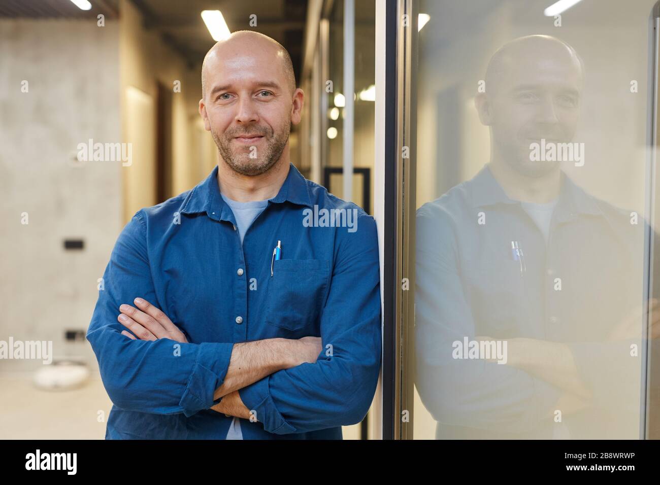 Waist up portrait of mature bald man smiling at camera while standing with arms crossed and posing confidently leaning against wall, copy space Stock Photo