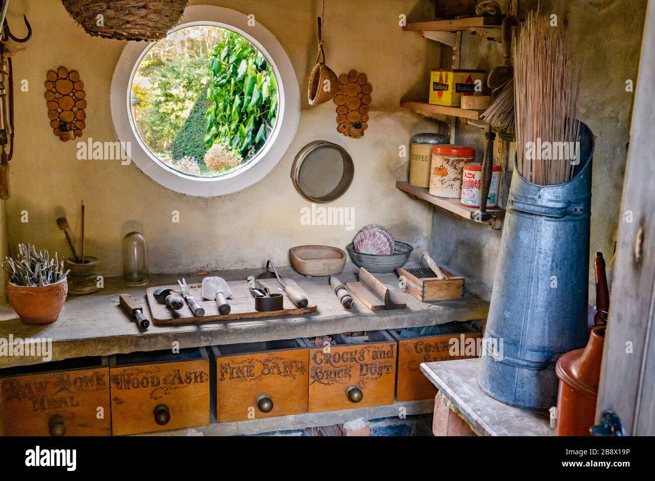 Old rustic potting shed interior, garden view through round window, gardening tools, workbench, retro containers & boxes - York Gate Garden,  Leeds UK Stock Photo