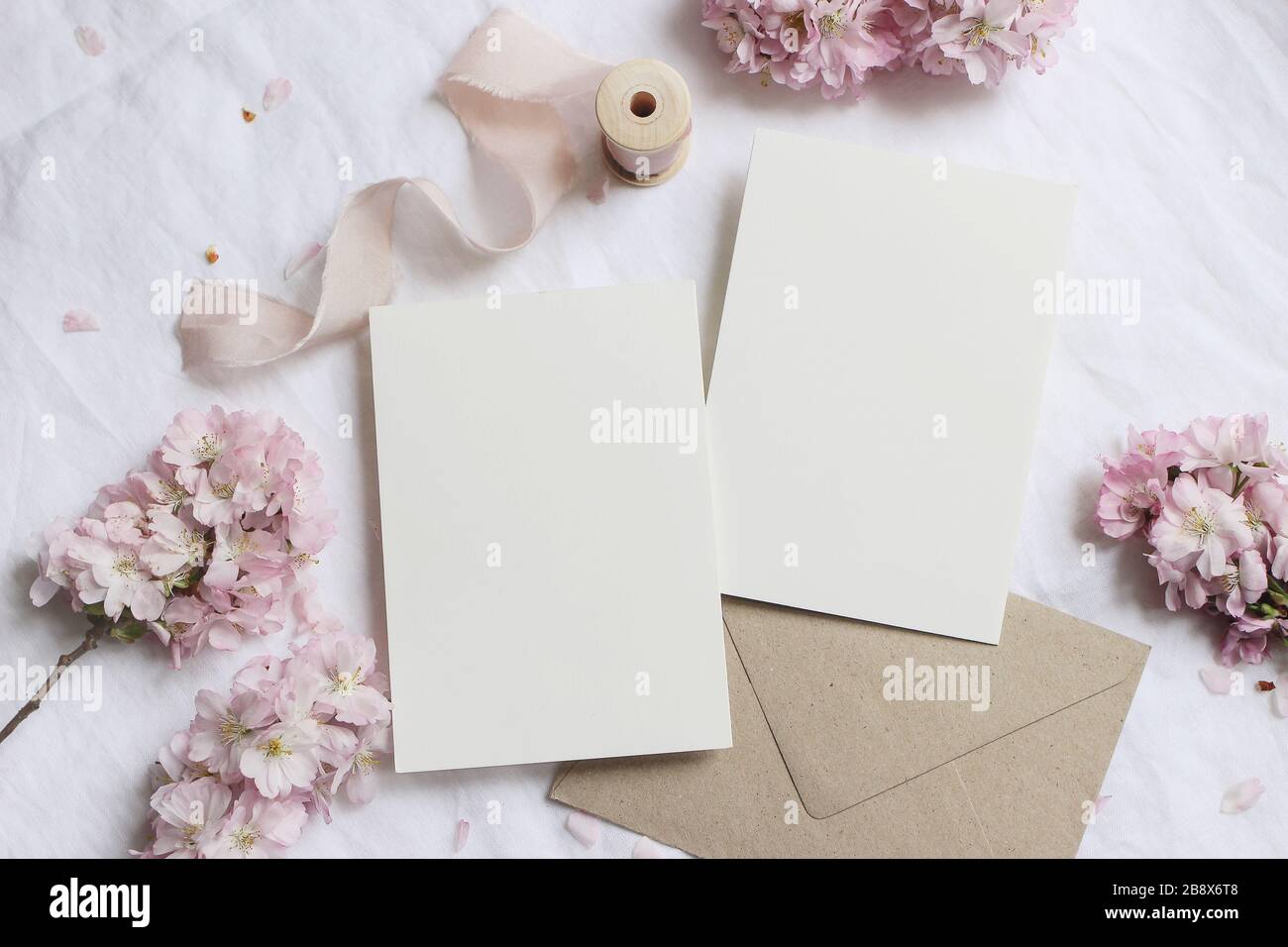 Wedding stationery mock-up scene. Blank greeting cards, envelope on linen tablecloth background with pink blossoming cherry tree branches and ribbon, Stock Photo