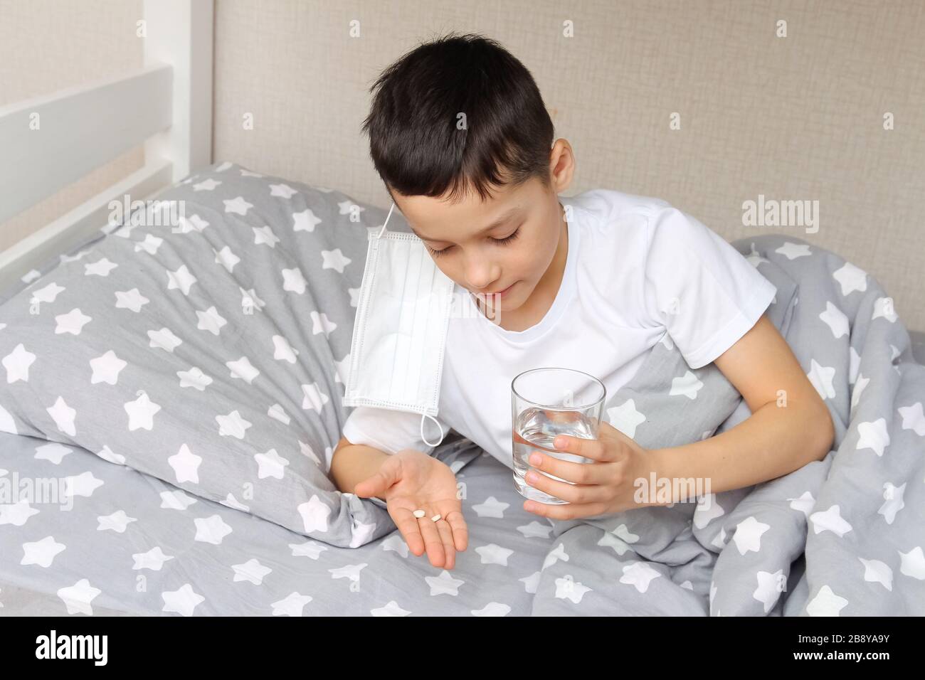 Caucasian boy with dark hair is lying in bed, holding pills and a glass of water, he is ill, taking medicine. The concept of home treatment. Stock Photo