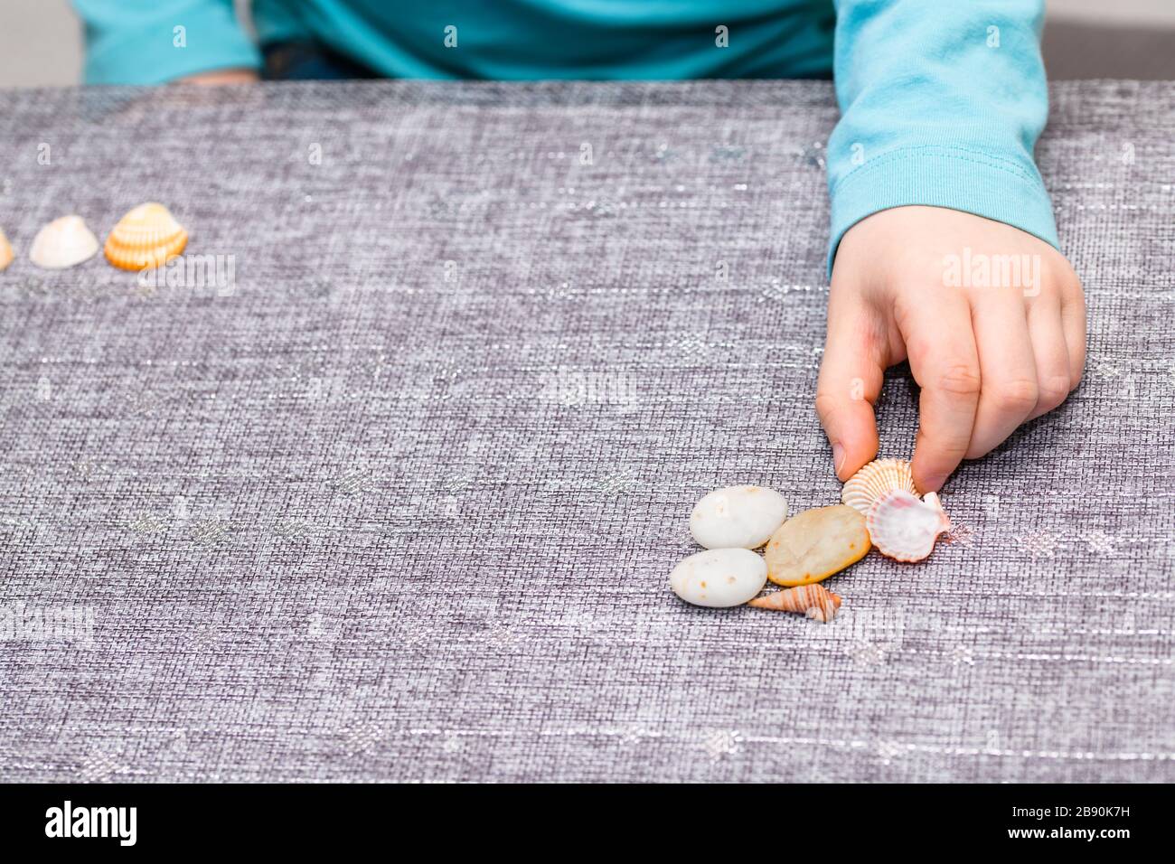 Five year old boy playing with a pile of seashells and pebbles. He has a turquoise shirt and picking up a seashell. Stock Photo