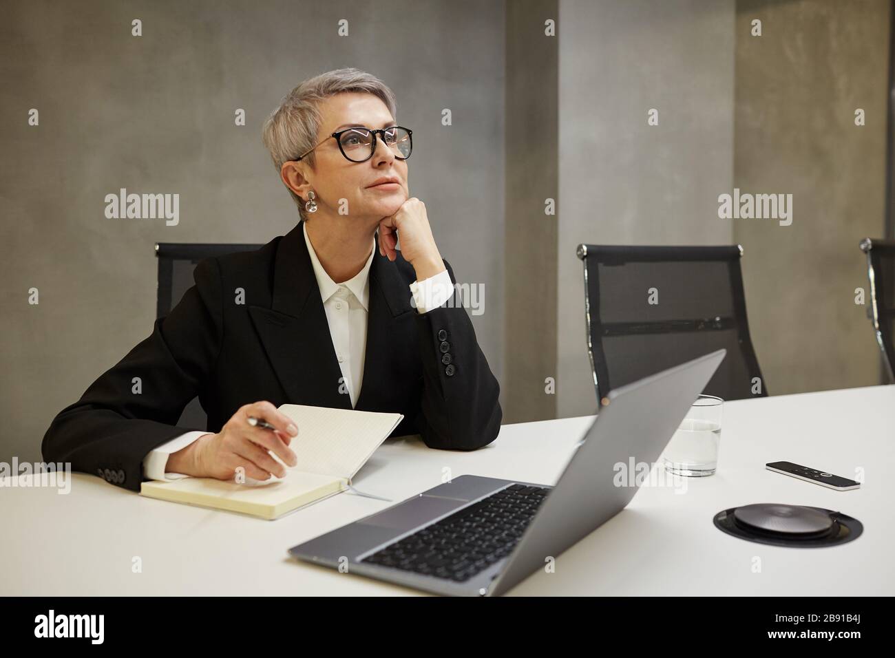 Minimal portrait of successful mature businesswoman looking away pensively while sitting against grey wall at workplace, copy space Stock Photo