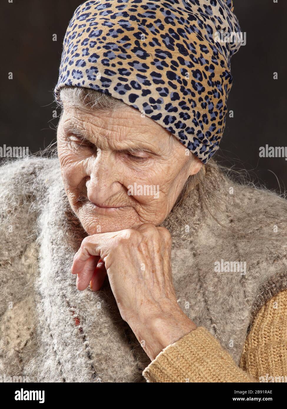 Close-up portrait of old woman with brooding look on dark background. She leaned her chin on her hand and deeply thought. Stock Photo